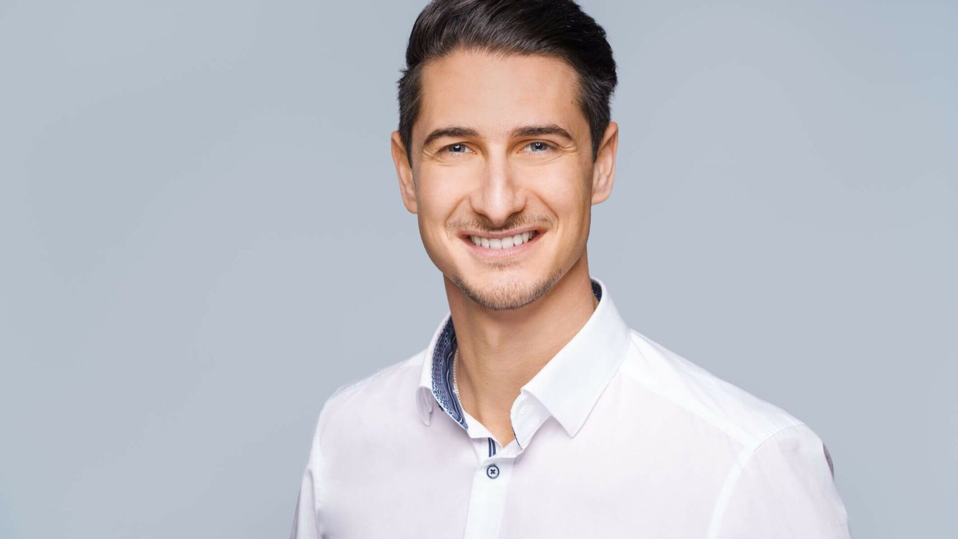 Meinkarrierestart.at Eine Person mit kurzen dunklen Haaren und hellem Bart lächelt in einem weißen Hemd mit Knöpfen vor einem grauen Hintergrund und fängt das ideale Foto für ein Bewerbungsschreiben ein.