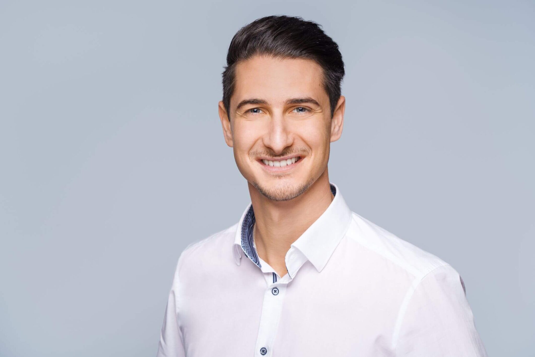 Meinkarrierestart.at Eine Person mit kurzen dunklen Haaren und hellem Bart lächelt in einem weißen Hemd mit Knöpfen vor einem grauen Hintergrund und fängt das ideale Foto für ein Bewerbungsschreiben ein.