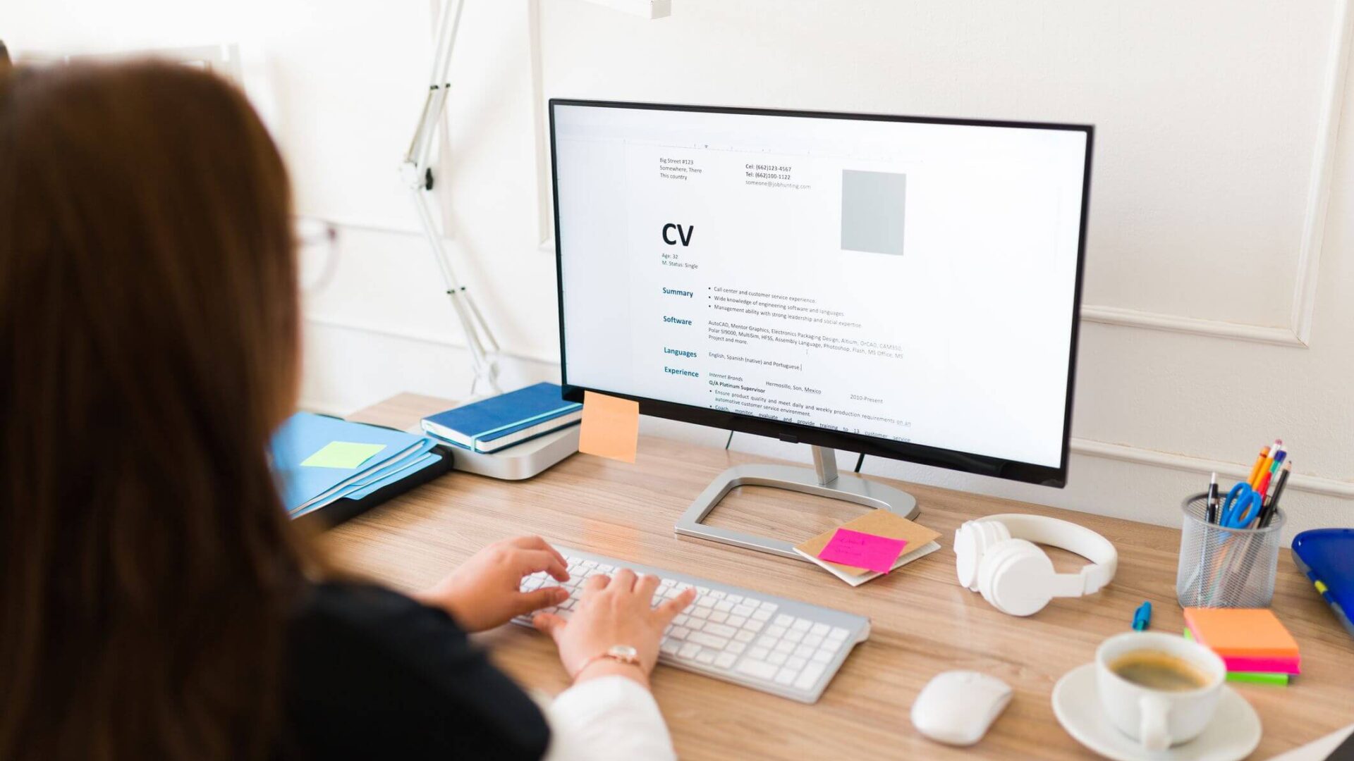 meinkarrierestart.at Eine Person sitzt an einem Schreibtisch und tippt auf einer Tastatur, während sie auf einem Computermonitor einen Lebenslauf ansieht, umgeben von Büromaterial, Kopfhörern und einer Tasse Kaffee.