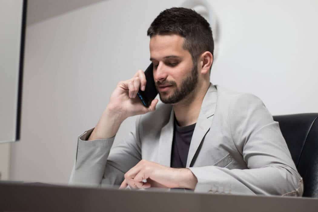 meinkarrierestart.at Ein Mann in einem hellgrauen Blazer sitzt an einem Schreibtisch, führt ein Telefoninterview, spricht mit seinem Smartphone und blickt auf die Oberfläche vor ihm.