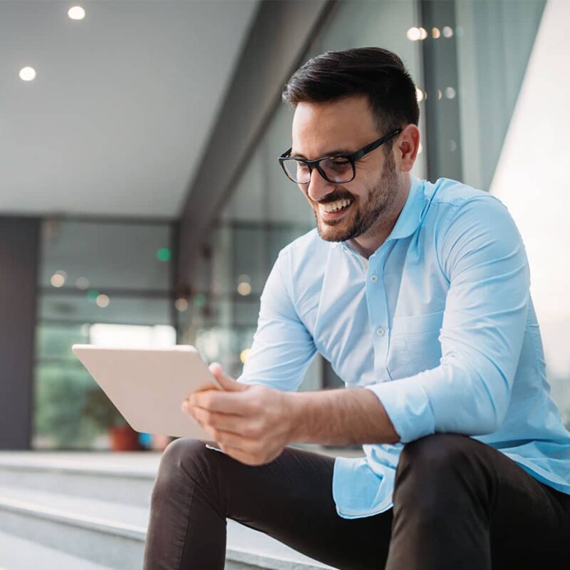 Meinkarrierestart.at Ein Mann mit Brille und Bart in einem hellblauen Hemd sitzt auf einer Treppe, lächelt und benutzt ein Tablet.