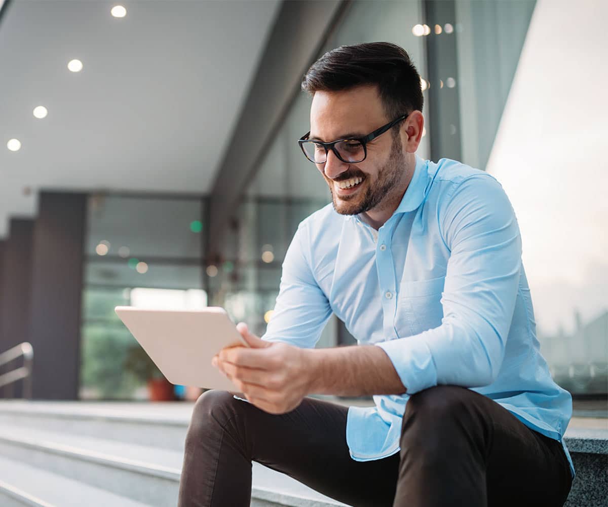 Meinkarrierestart.at Ein Mann mit Brille und Bart in einem hellblauen Hemd sitzt auf einer Treppe, lächelt und benutzt ein Tablet.