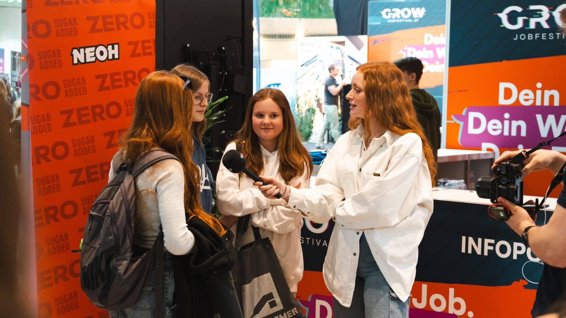 meinkarrierestart.at Eine Frau mit einem Mikrofon interviewt drei Mädchen bei einer Indoor-Veranstaltung mit Informationsständen und Bannern im Hintergrund.