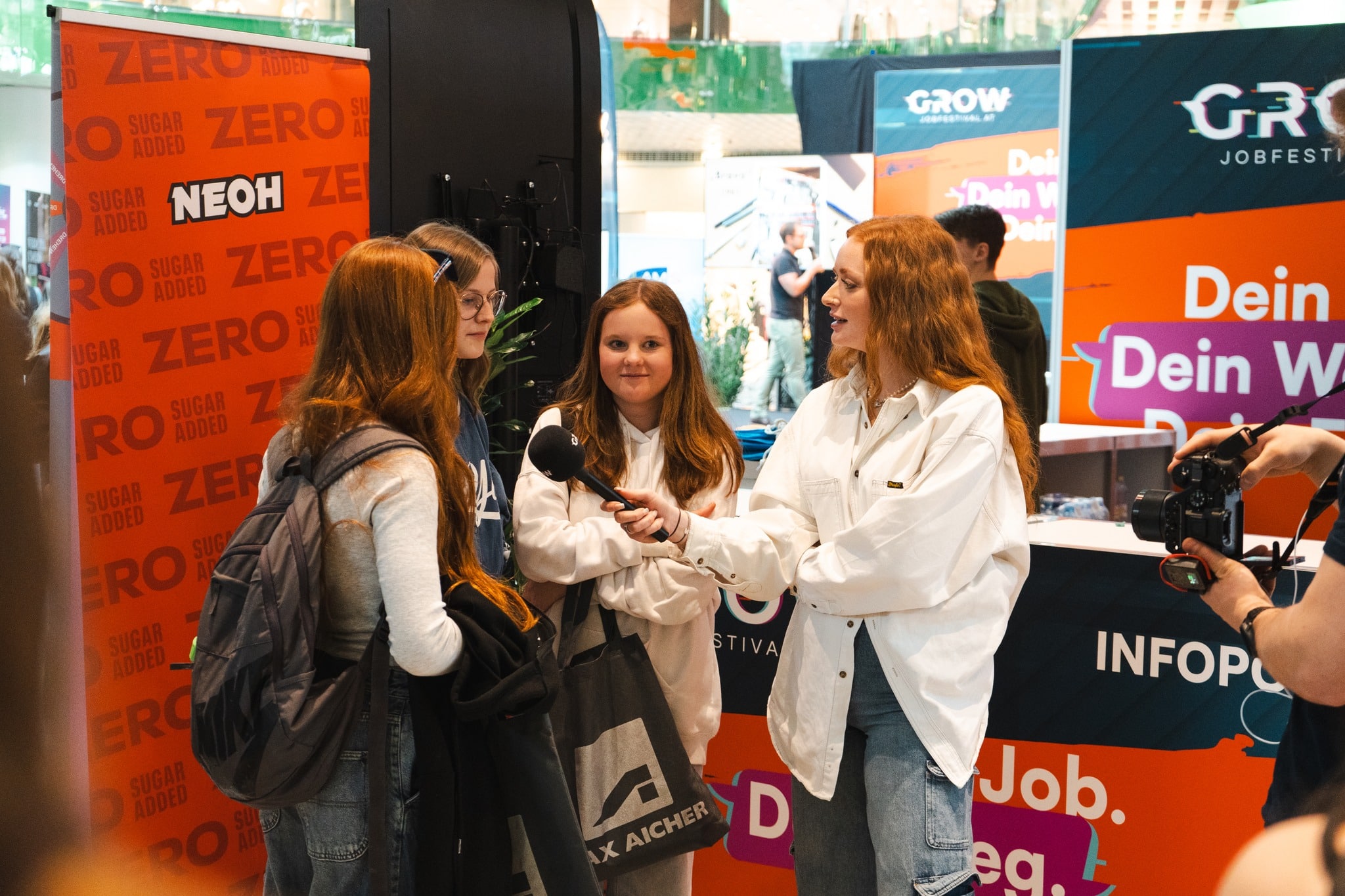 meinkarrierestart.at Eine Frau mit einem Mikrofon interviewt drei Mädchen bei einer Indoor-Veranstaltung mit Informationsständen und Bannern im Hintergrund.