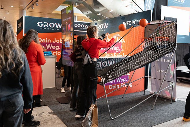 meinkarrierestart.at An einem Messestand auf einem Jobfestival mit orange-weißen Werbebannern im Hintergrund spielen Leute ein Indoor-Basketball-Arcade-Spiel.
