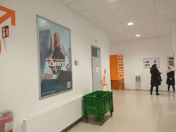 meinkarrierestart.at Ein Flur mit weißen Wänden, einem grünen Plastikwagen, einem großen Plakat und zwei Personen, die neben einer orangefarbenen Tür mit der Aufschrift „PORTIER“ stehen.