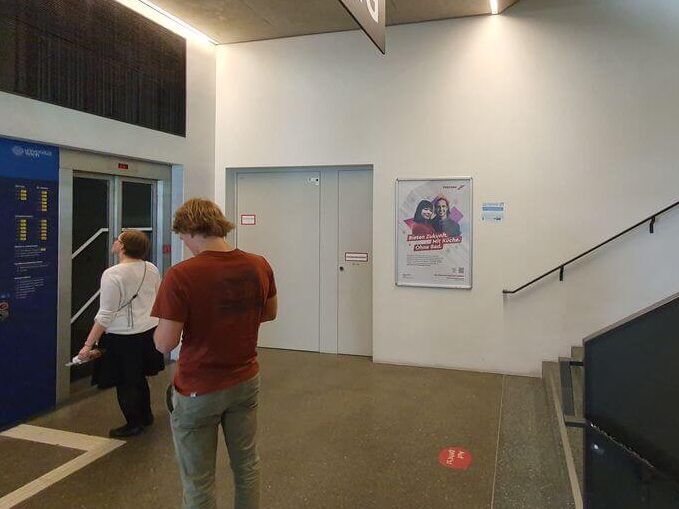 meinkarrierestart.at Ein Mann steht in der Lobby eines Gebäudes neben einem Aufzug und einem Wandplakat. Rechts befindet sich eine Treppe, und von der Decke hängt ein großes Schild mit der Aufschrift „EG“.