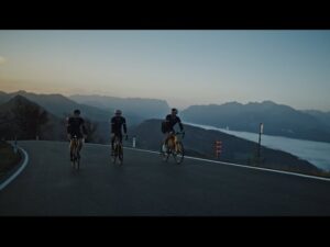 meinkarrierestart.at Drei Radfahrer fahren im Morgengrauen eine asphaltierte Straße bergauf, im Hintergrund sind Berge und ein nebelverhangenes Tal zu sehen.