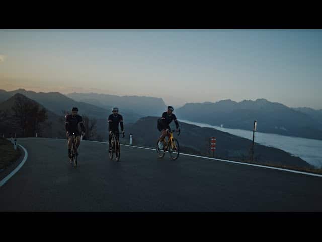 meinkarrierestart.at Drei Radfahrer fahren im Morgengrauen eine asphaltierte Straße bergauf, im Hintergrund sind Berge und ein nebelverhangenes Tal zu sehen.