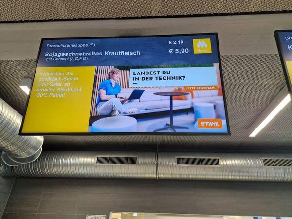 meinkarrierestart.at Auf einer digitalen Menütafel werden die Menüoptionen und Preise auf Deutsch angezeigt, außerdem gibt es eine Anzeige mit einer Frau mit Laptop und dem Slogan „Landest du in der Technik?“.
