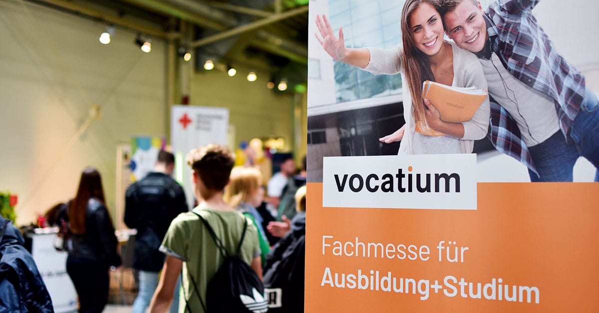 meinkarrierestart.at Eine Berufsmesse, bei der Besucher an einem Schild mit der Aufschrift „vocatium Fachmesse für Ausbildung+Studium“ vorbeigehen, auf dem zwei lächelnde junge Erwachsene mit Büchern in der Hand zu sehen sind.