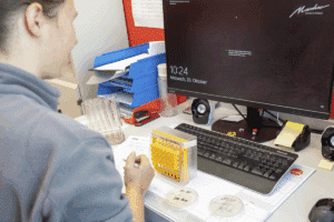 meinkarrierestart.at Eine Person an einem Schreibtisch mit Laborgeräten, Petrischalen und einem Computer, der Uhrzeit und Datum anzeigt, arbeitet in einem Büro oder Labor.
