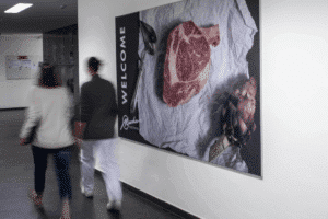 meinkarrierestart.at Zwei Personen gehen an einem Wandplakat vorbei, auf dem ein rohes Ribeye-Steak, eine Artischocke und in großen Buchstaben das Wort „WILLKOMMEN“ abgebildet sind.