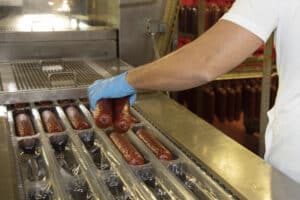 meinkarrierestart.at Eine Person mit blauen Handschuhen legt an einer industriellen Lebensmittelproduktionsstraße Würstchen in Verpackungsschalen aus Kunststoff.
