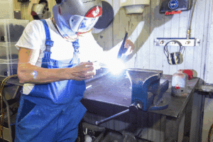 meinkarrierestart.at Person mit Schweißhelm und blauem Overall schweißt Metall an einer Werkbank in einer Werkstatt, helle Funken sind sichtbar.