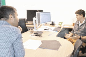 meinkarrierestart.at Zwei Männer sitzen sich an einem Schreibtisch in einem Büro gegenüber und unterhalten sich. Auf dem Schreibtisch liegen Papiere und ein Computerbildschirm.