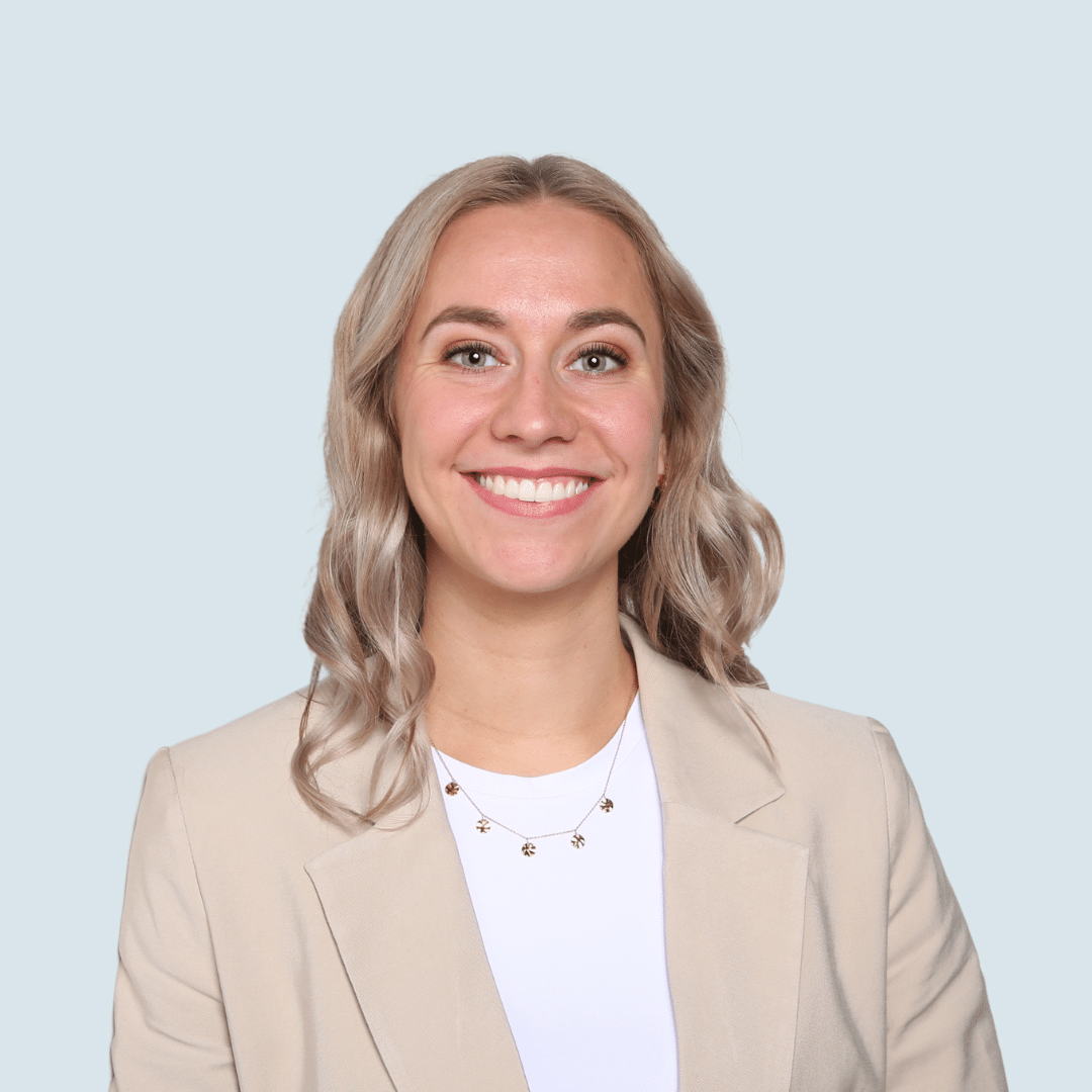 meinkarrierestart.at Eine Frau mit blondem, welligem Haar, einem beigen Blazer und einem weißen Oberteil lächelt vor einem hellgrauen Hintergrund in die Kamera.